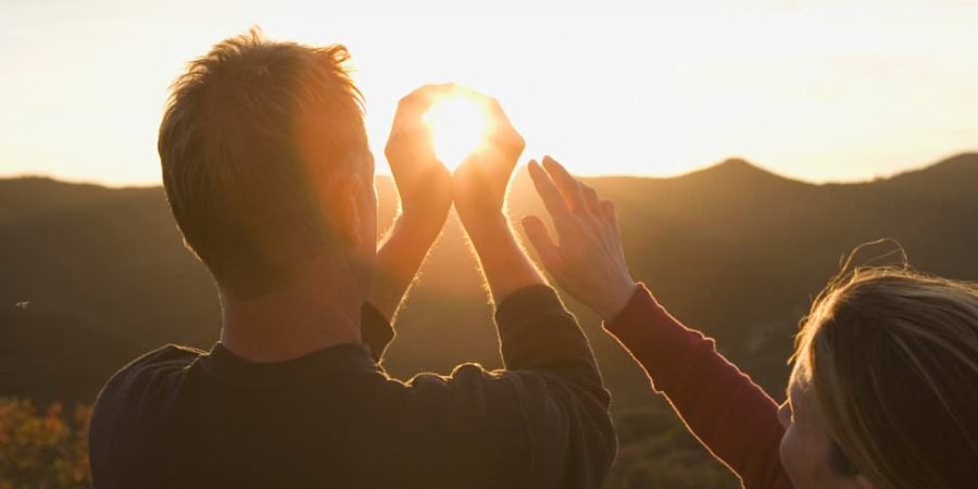 Muž a žena pozorují západ slunce, přičemž muž dělá gesto, jako by rukama držel slunce.
