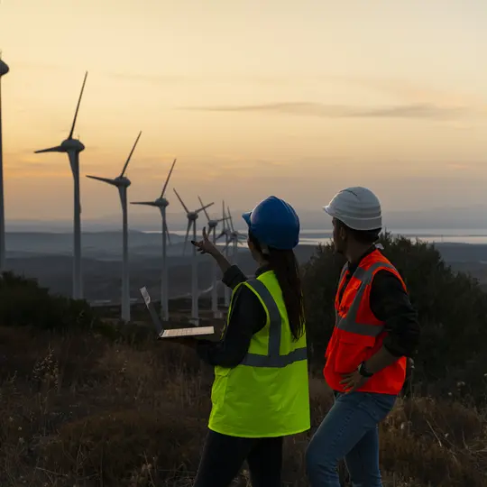 Two individuals in safety vests standing beside wind turbines, representing smart grid solutions for electric utilities.