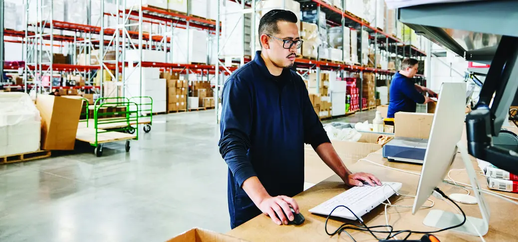 Male warehouse worker checking orders at computer work station in warehouse