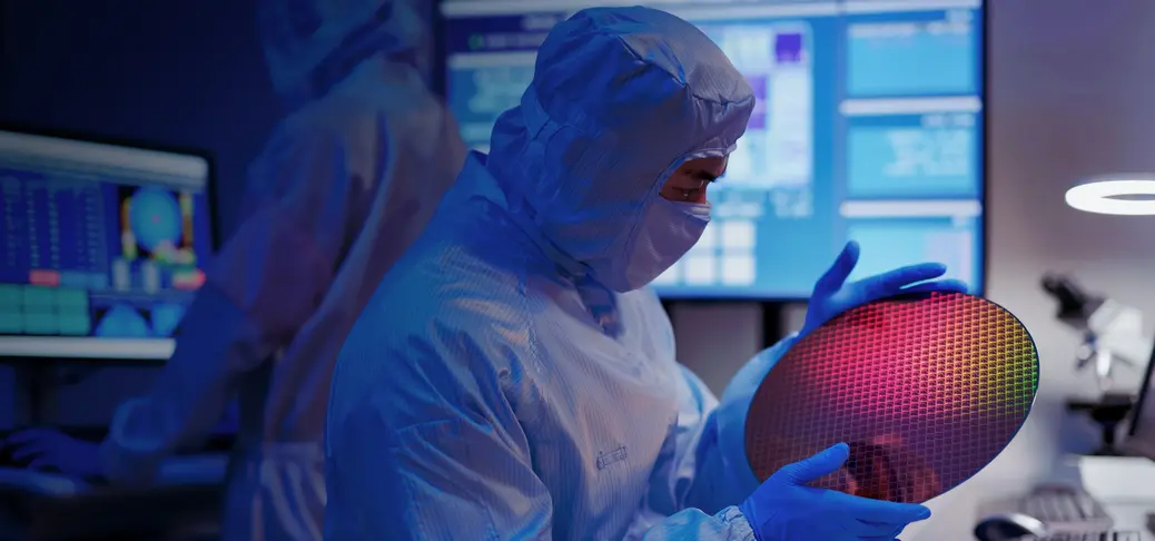 Technician in sterile coverall holds wafer that reflects many different colours with gloves and check it at semiconductor manufacturing plant