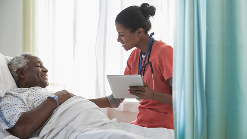 A nurse holding a tablet with a patient lying in a bed