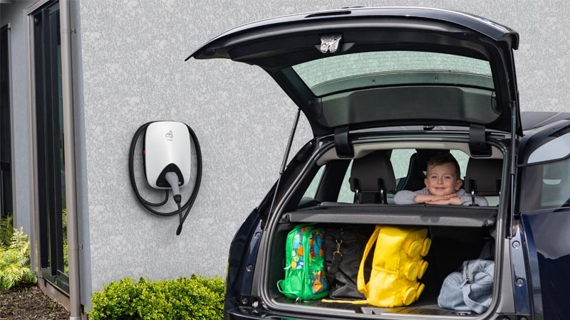 Car with boot open showing bags and a child in the back seat. Electric vehicle charger on the house wall in the background.