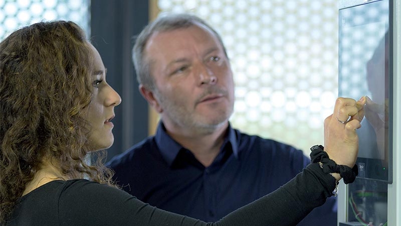 A woman using a touch screen in a server room while talking to a man by her side