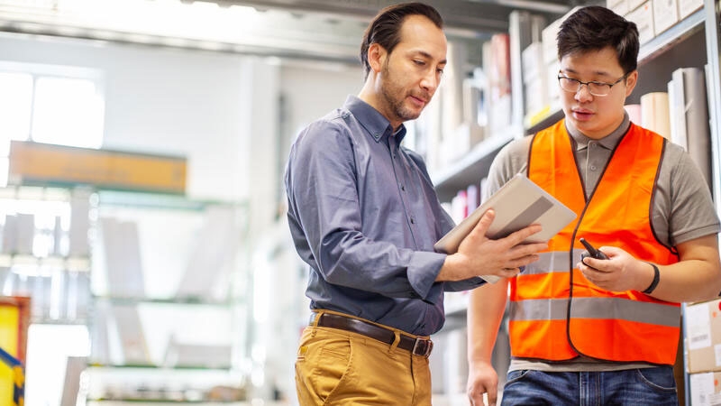 Warehouse supervisor showing digital tablet to the worker in factory storage room. Warehouse workers working together, looking at tablet pc and discussing.