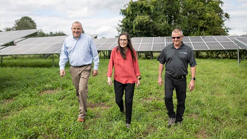 A group of three people walk on a green field with sun panels behind them