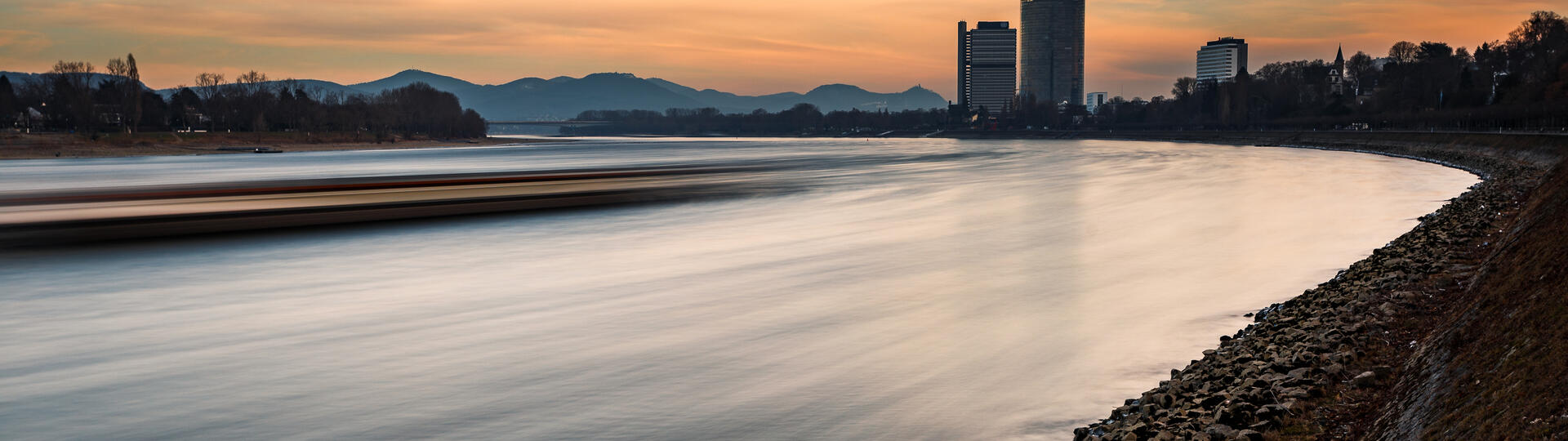 The riverside of the rhine river in Bonn with a moving ship in the front and the dhl-tower in the background during sunset.